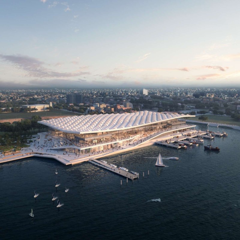Aerial view of a modern waterfront building with a wavy roof and boats docked nearby.