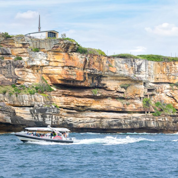Speedboat on blue ocean near rocky, colorful cliff with small building on top.
