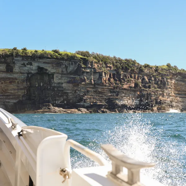 Boat sailing near rocky cliff coastline with splashing water under a clear blue sky.