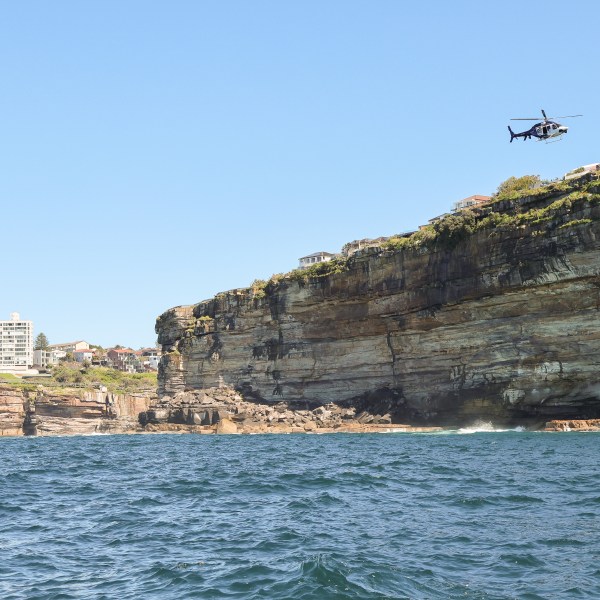Helicopter flying over coastal cliff with ocean and buildings in the background on a Sydney offshore cruise.