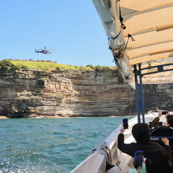 Boat passengers take photos of a helicopter flying near a rocky cliff under a clear blue sky off Sydney Harbour.