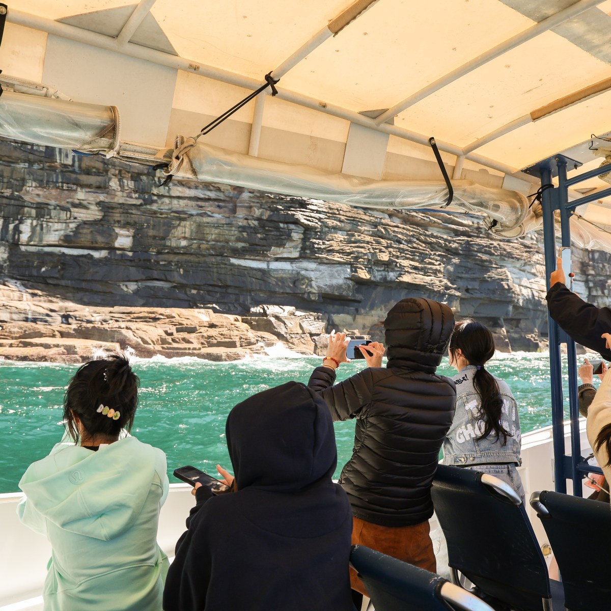 People on a boat taking photos of rocky cliffs over green water.