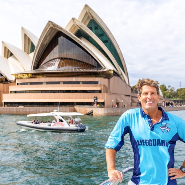 Person in blue lifeguard shirt standing in front of Sydney Opera House with a boat on the water.