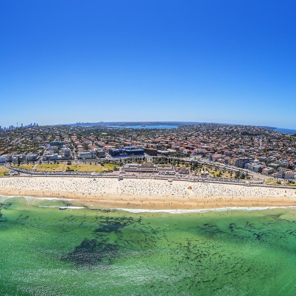 Aerial view of a crowded beach with a city and ocean backdrop under a clear blue sky.