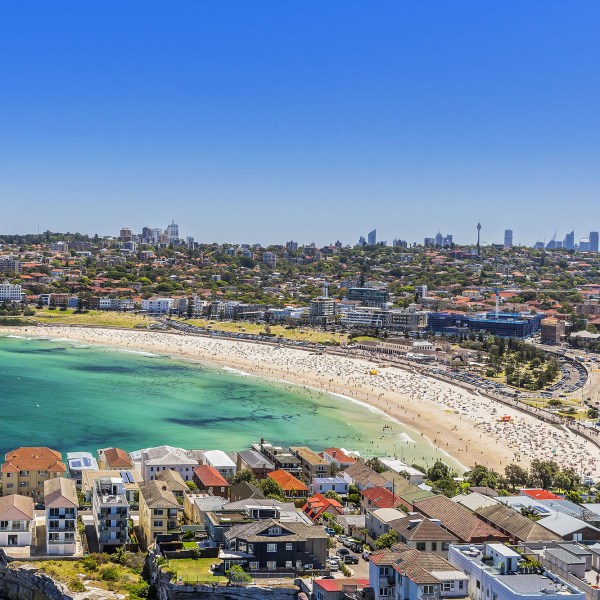 Aerial view of a crowded beach with a curved shoreline, houses, and distant city skyline.