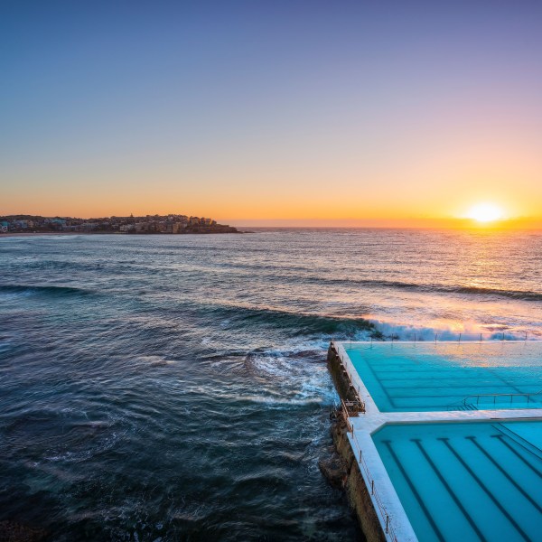 Coastal pool at sunrise with ocean waves and distant buildings.