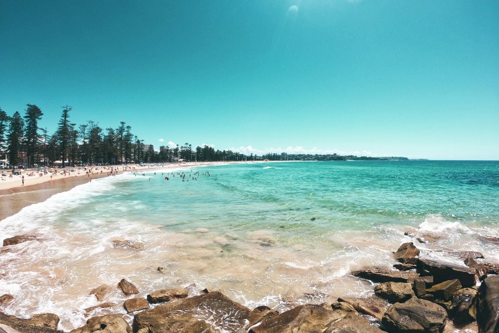 Sunny beach with turquoise water, rocky shore, and people swimming, with trees lining the distant coastline.