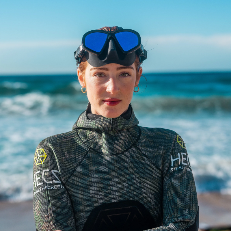 Person in wetsuit and dive mask stands on beach with waves in background.