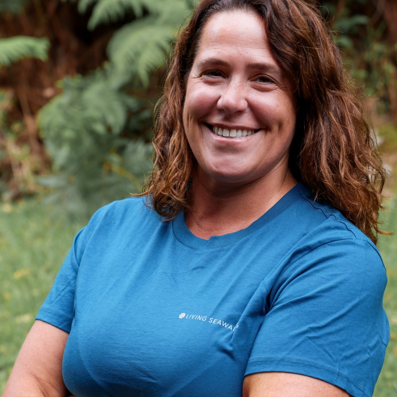 Smiling person with curly hair in a blue shirt stands cross-armed outdoors.