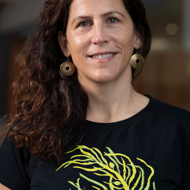 Smiling woman with long hair, wearing large round earrings and a black shirt with green leaf pattern.
