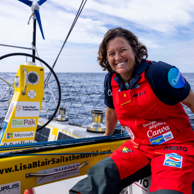 Person smiling on a sailboat, wearing a red sailing suit, with logos on clothing and equipment.
