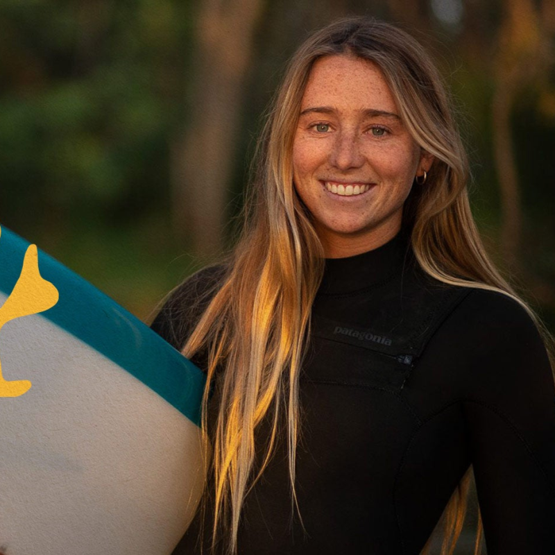 Woman in wetsuit holding a surfboard, smiling against a blurred natural background.