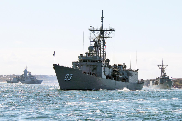 Three naval ships sail in formation on a sunny day with clear skies.