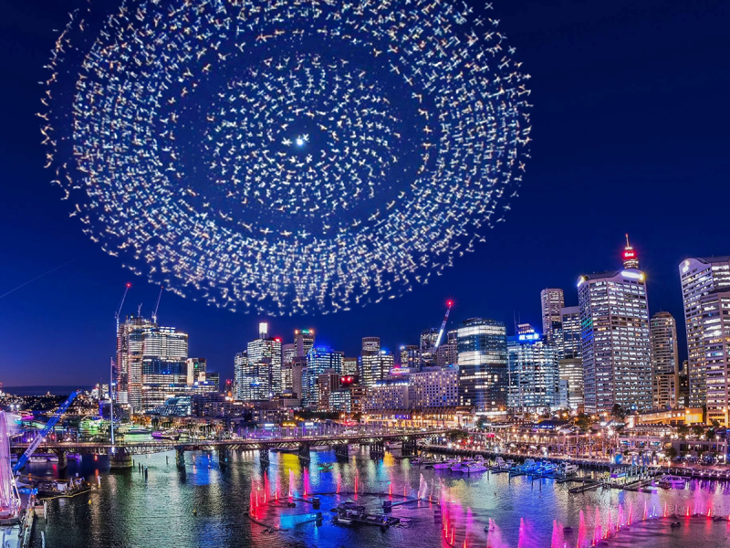Cityscape at night with a large circular drone light display in the sky.