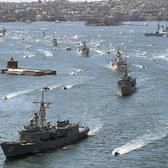 Aerial view of naval ships and boats sailing in a harbor, with buildings in the background.