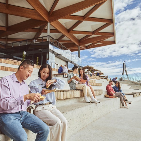 People sitting on outdoor steps, eating and talking under a modern wooden canopy.