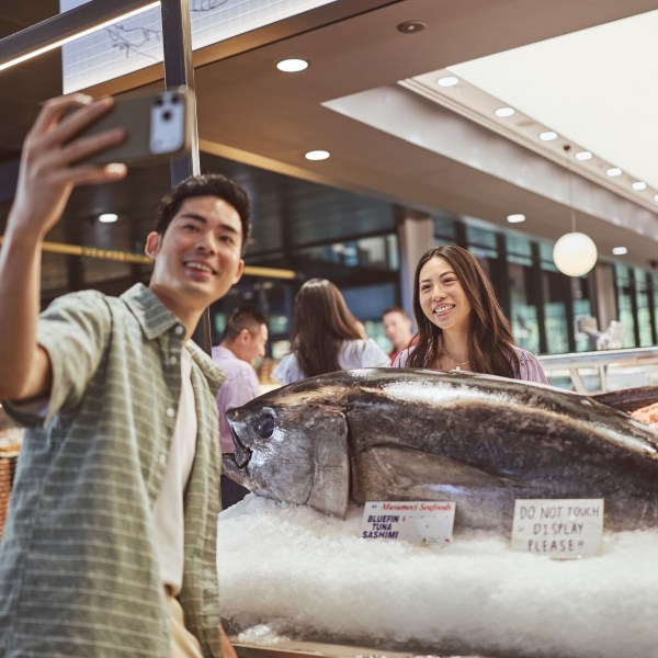 Two people taking a selfie with a large fish on display in a market.