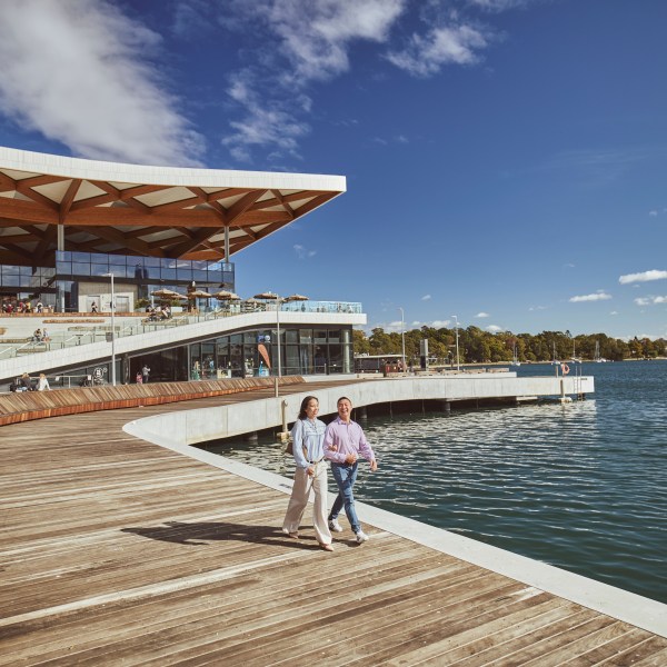 Two people walking on a boardwalk by a modern waterfront building.
